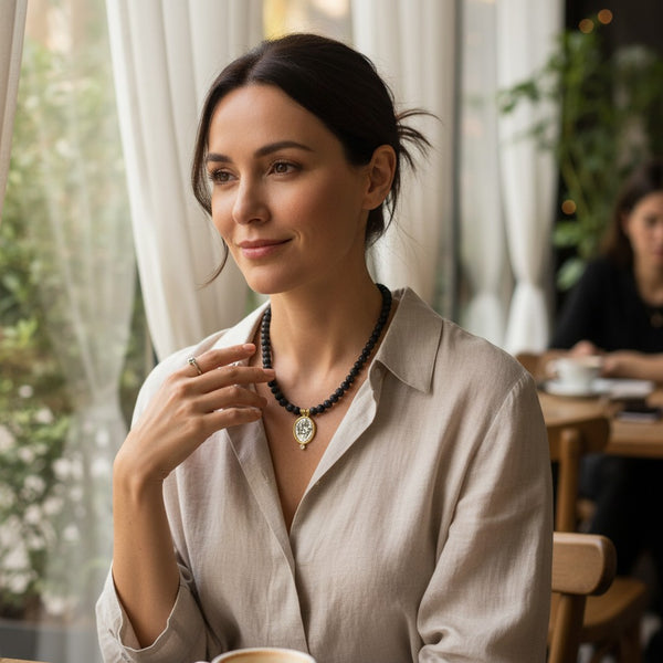 Woman wearing an elephantina.gr black beaded necklace with a gold-framed oval pendant in a cafe setting
