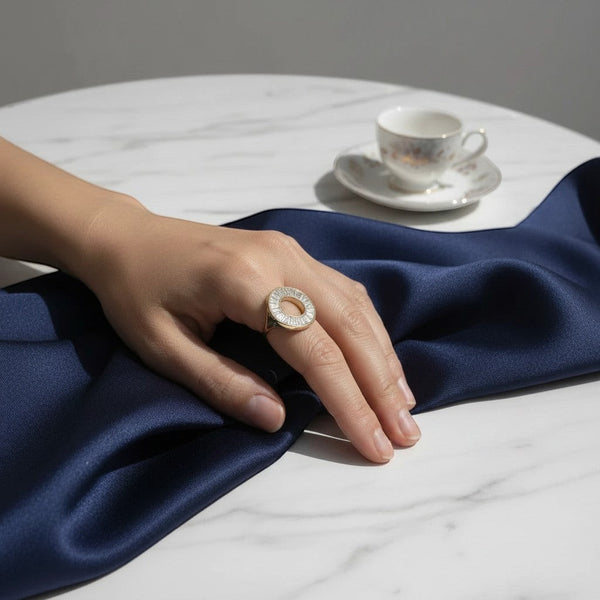 elephantina.gr gold-tone circular statement ring with baguette-cut crystals on a hand over navy silk fabric.