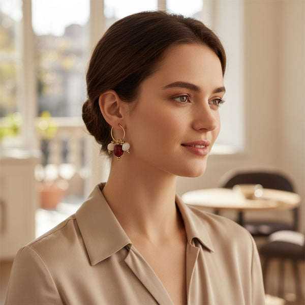 A woman wearing elephantina.gr gold hoop earrings with red and white beads in a bright, elegant indoor setting.