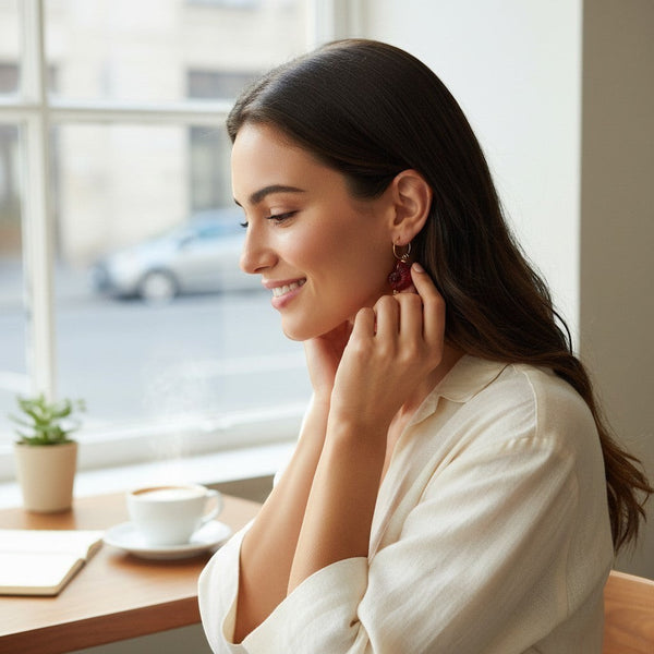 A woman wearing elephantina.gr gold hoop earrings with red floral charms while sitting in a bright cafe.