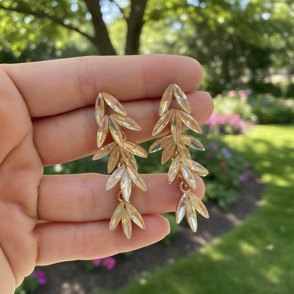 elephantina.gr gold leaf-shaped crystal drop earrings held in hand against a blurred garden background