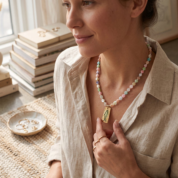 elephantina.gr necklace featuring pastel beads and a rectangular gold pendant with a green fish design on a woman