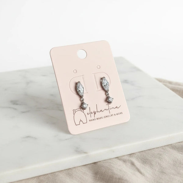 Κρεμαστά σκουλαρίκια elephantina.gr από ορείχαλκο σε ασημί χρώμα με λευκά κρύσταλλα σε σχήμα νυχάκι πάνω σε κάρτα.