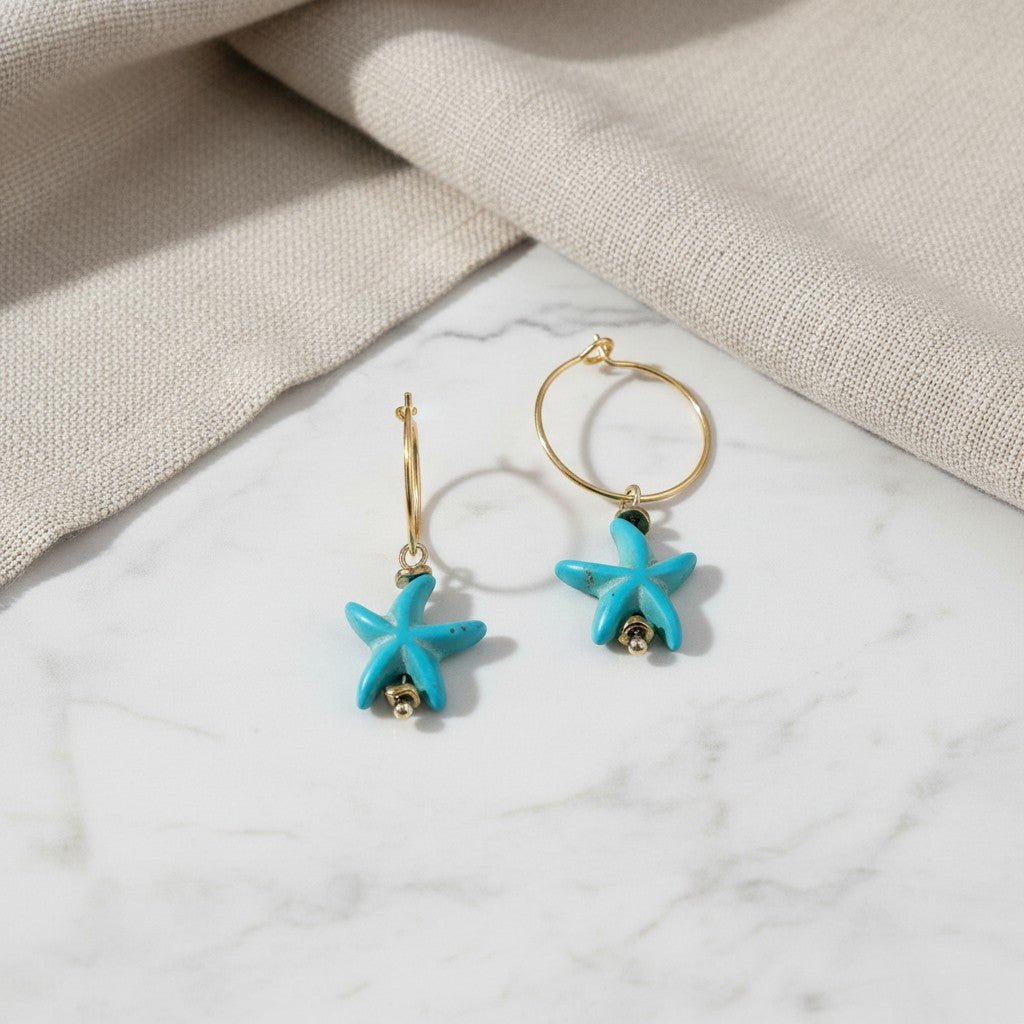 elephantina.gr gold hoop earrings featuring turquoise starfish charms on a white marble surface with beige fabric