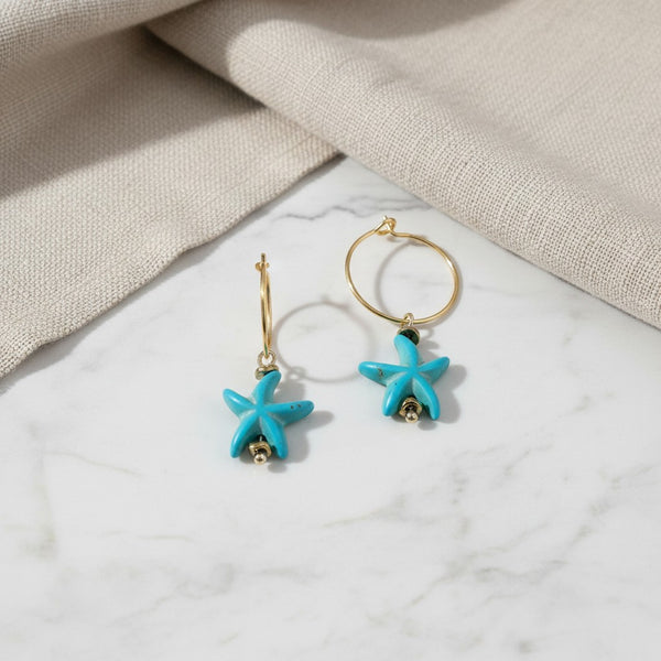 elephantina.gr gold hoop earrings featuring turquoise starfish charms on a white marble surface with beige fabric