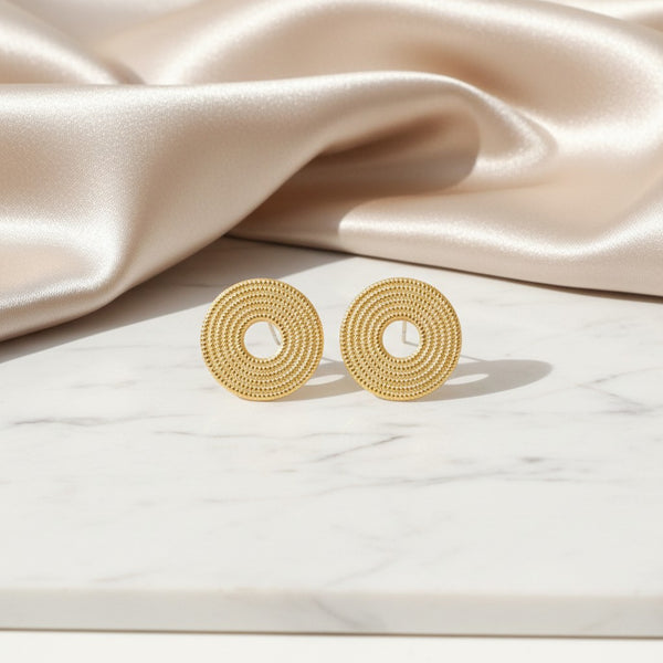 elephantina.gr 24K gold-plated brass round stud earrings with textured detail on a marble surface and silk fabric.