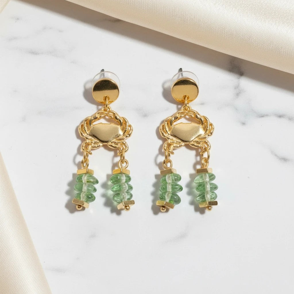 elephantina.gr gold-plated stud earrings with crab charms and green glass beads on a white marble background