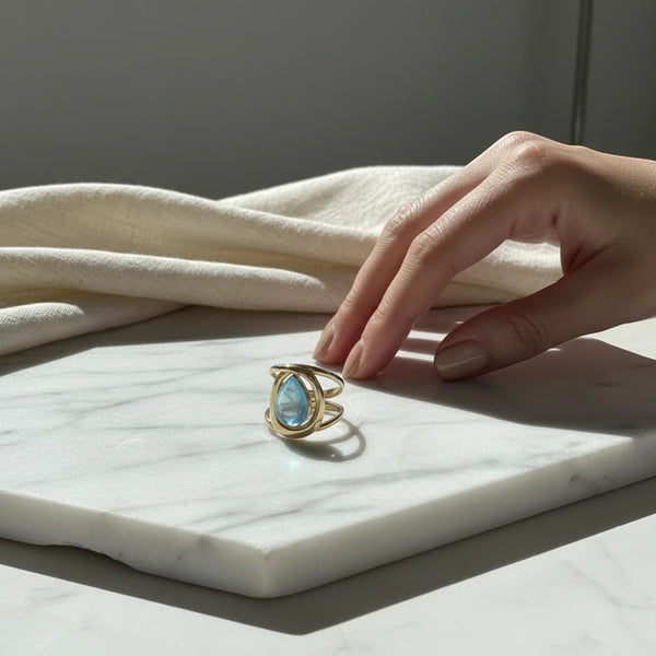 elephantina.gr gold statement ring with a teardrop blue stone on a marble surface with a hand nearby.