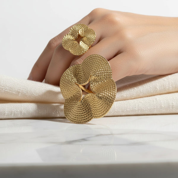 elephantina.gr gold-tone statement ring with a sculptural, textured floral design worn on a hand against white fabric.
