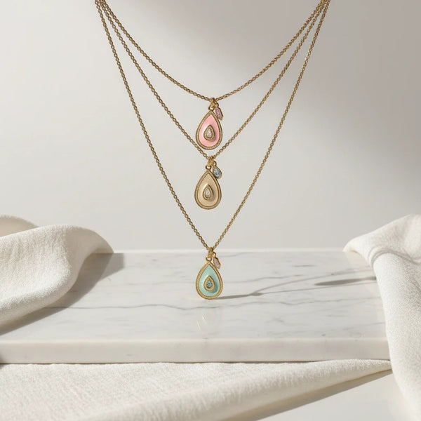elephantina.gr layered gold necklaces with pastel teardrop enamel pendants in pink, cream, and blue with glitter detail.