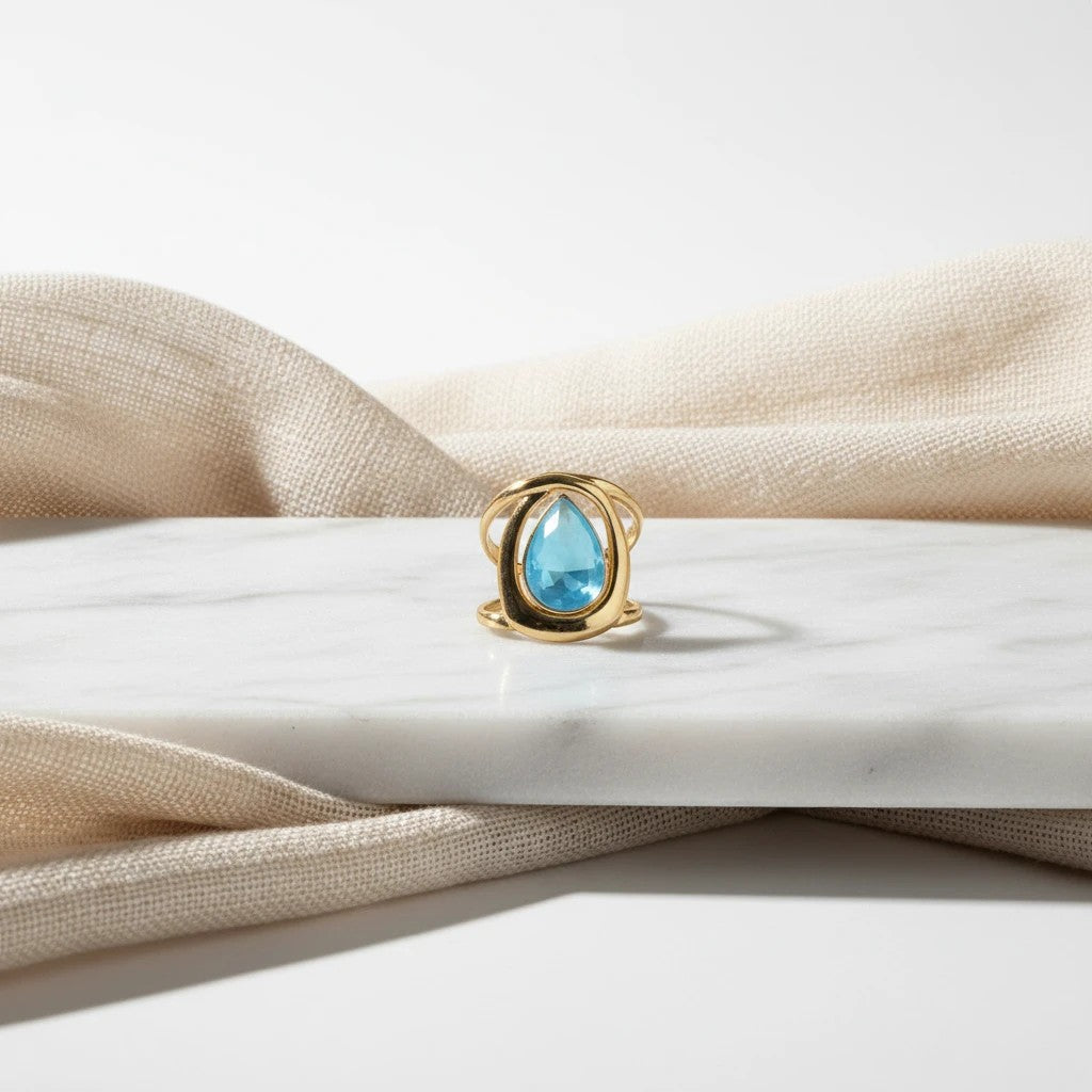 elephantina.gr statement ring with a teardrop blue stone in a polished gold-tone open-frame setting on marble.
