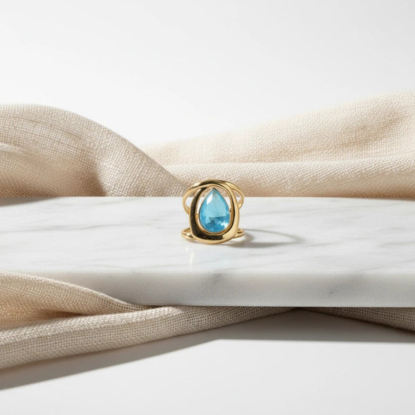 elephantina.gr statement ring with a teardrop blue stone in a polished gold-tone open-frame setting on marble.