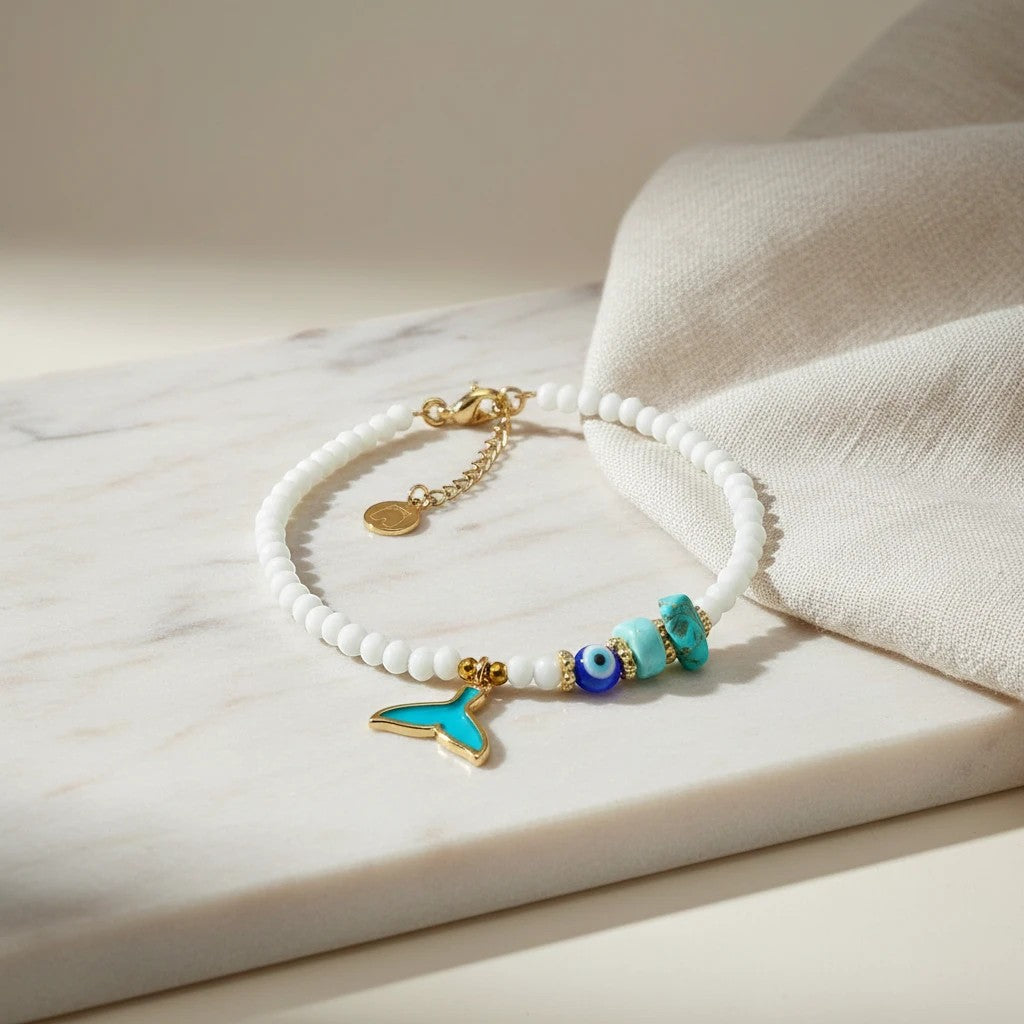 White beaded anklet with gold whale tail charm, turquoise beads, and evil eye charm. 24K gold plated.