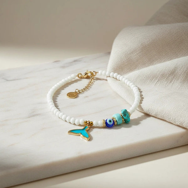 White beaded anklet with gold whale tail charm, turquoise beads, and evil eye charm. 24K gold plated.