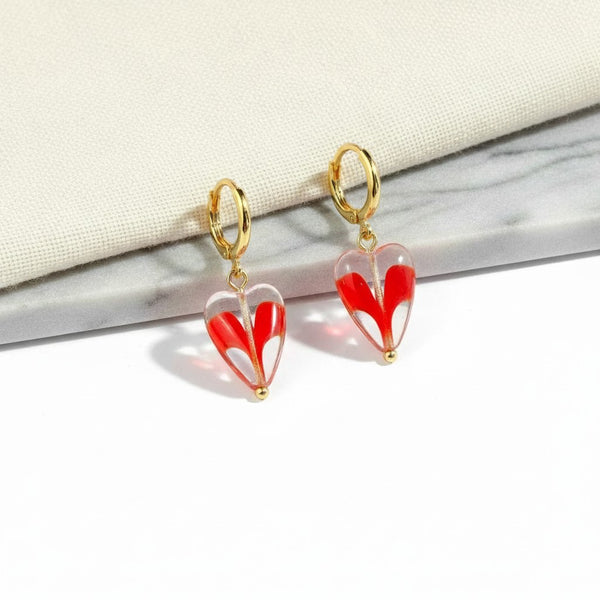 elephantina.gr handmade brass hoop earrings with dangling red heart charms on a marble and fabric background.