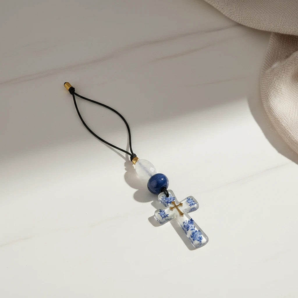 Decorative Christian cross ornament with blue and white floral pattern, clear material, gold accent, and blue/white beads on black cord.