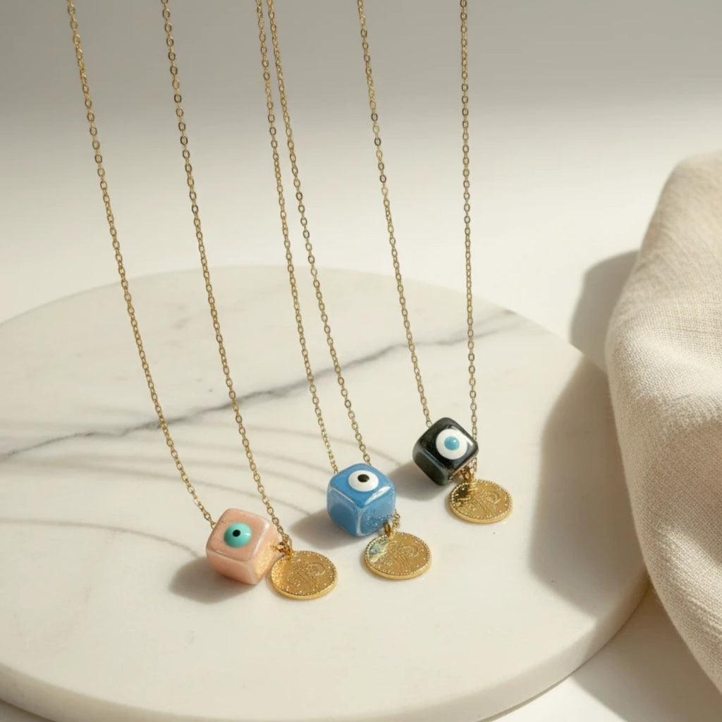 Three elephantina.gr necklaces with gold chains, ceramic evil eye cubes in pink, blue, and black, and gold coin pendants.