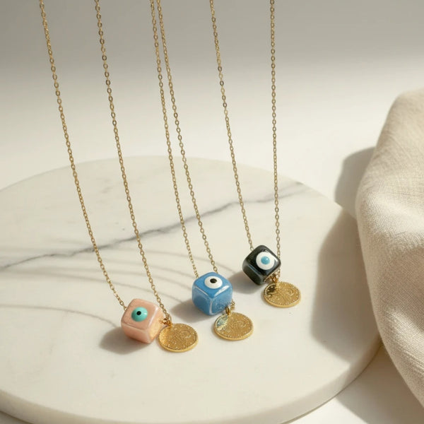 Three elephantina.gr necklaces with gold chains, ceramic evil eye cubes in pink, blue, and black, and gold coin pendants.